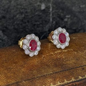Gold stud earrings with oval red rubies surrounded by a diamond halo on a rustic surface
