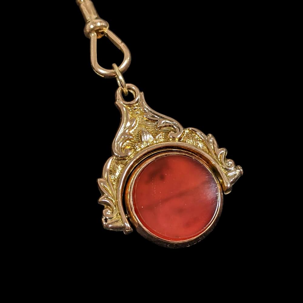 Ornate gold pendant with a red circular cabochon stone on a chain against a black background