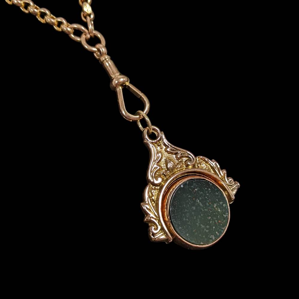 Gold pendant necklace with an ornate leaf-pattern frame around a dark green circular stone on a matching chain.