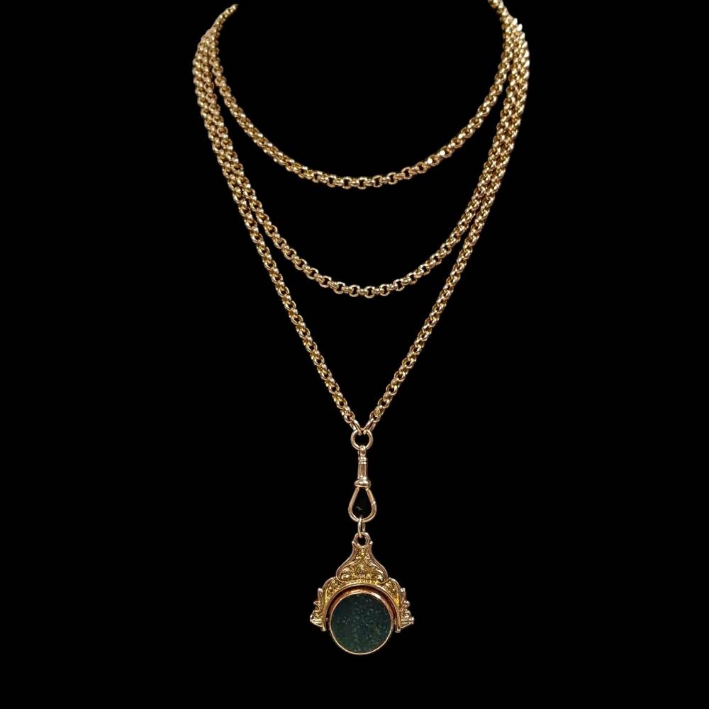 Gold layered chain necklace with an ornate pendant featuring a green circular stone on a black background.