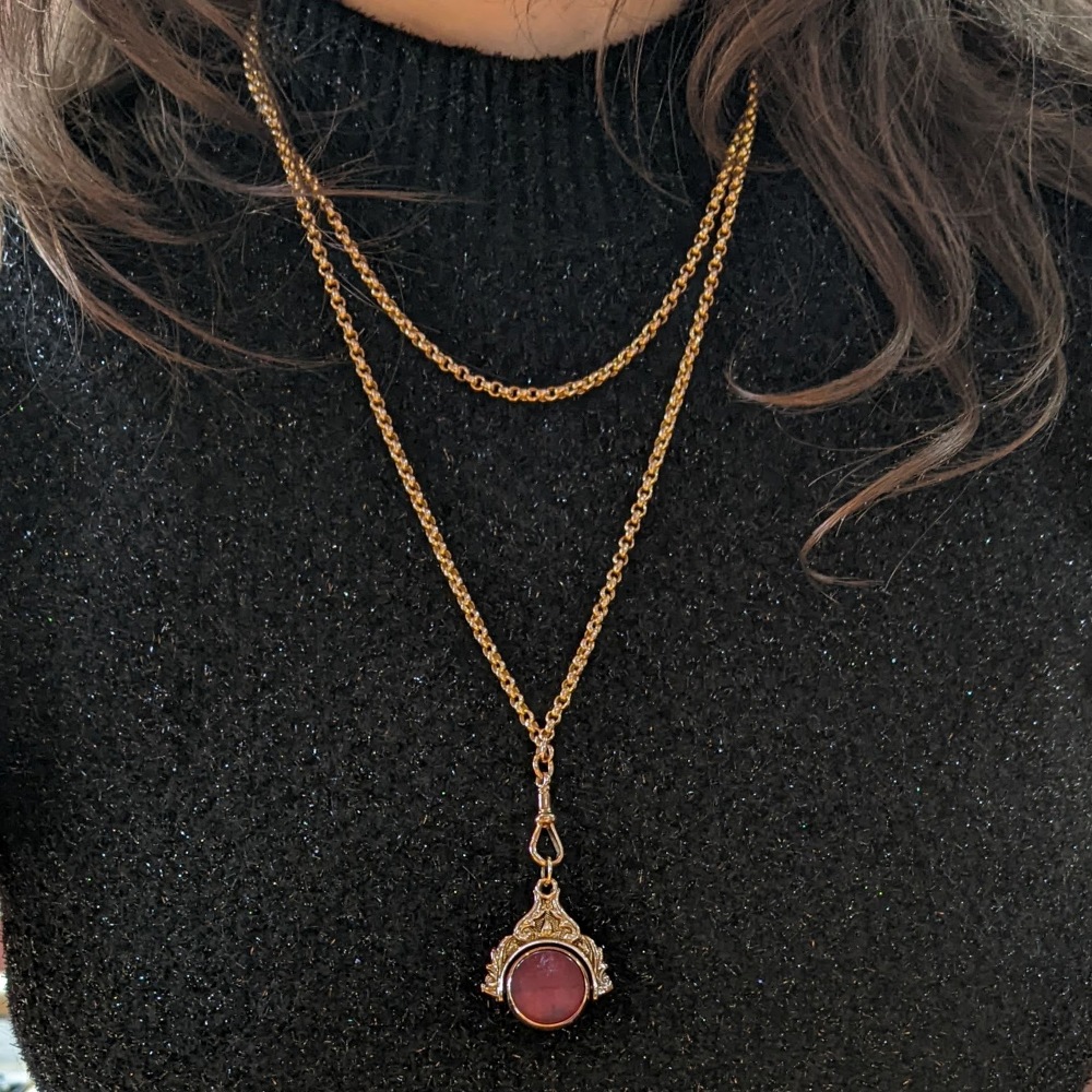 Gold layered necklaces with a pendant featuring a pink circular gemstone on a black glittery sweater.