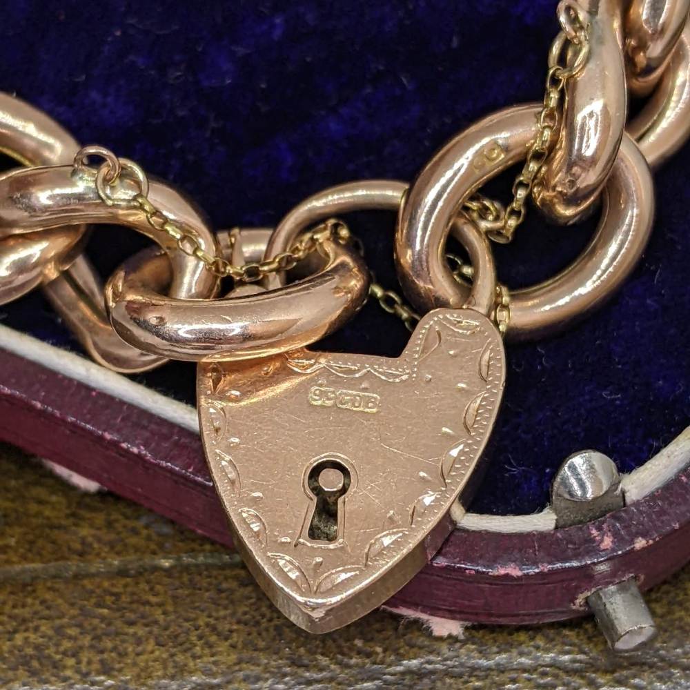 Heart-shaped rose-gold padlock with keyhole attached to chunky gold chain on a dark blue velvet and leather surface, jewelry-like.