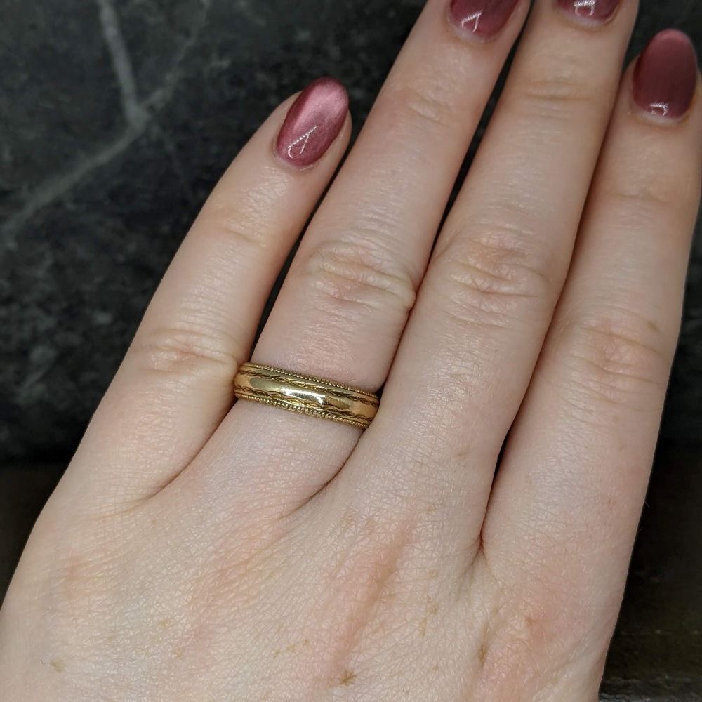 Close-up of a pale hand wearing a gold engraved band on the ring finger, dark burgundy nails visible.