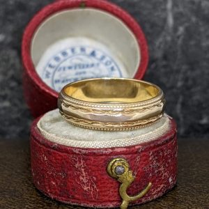 Gold ring with intricate braided and dotted engravings resting in a red velvet ring box.