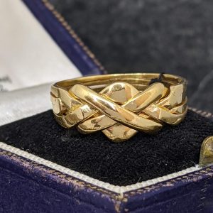 Gold braided ring with a knot-like design in a dark blue jewelry box, showcasing polished surfaces.