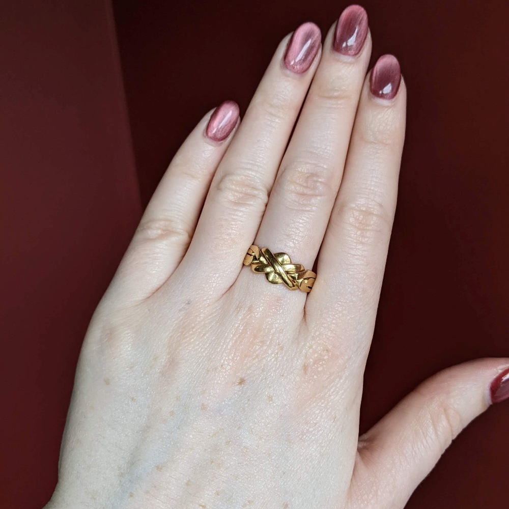 Close-up of a left hand with a gold braided band ring on the ring finger and pink manicured nails.