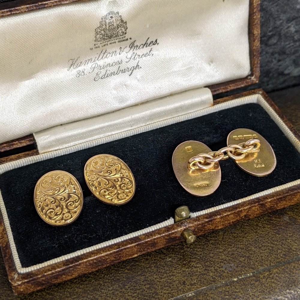 Open vintage leather jewelry box with black velvet interior, showing ornate gold cufflinks linked by a chain and two engraved gold studs; satin lining bears a script logo (Hamilton & Inches).