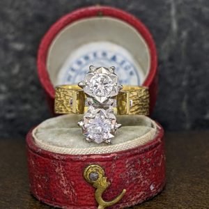 Two diamond rings stacked together in a red velvet ring box, one with a textured gold band and two prominent round stones