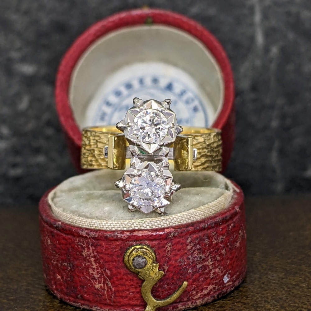 Two diamond rings stacked together in a red velvet ring box, one with a textured gold band and two prominent round stones