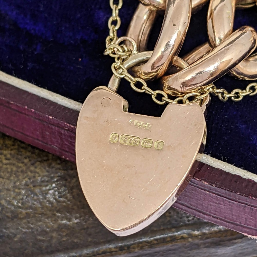 Close-up of a rose-gold heart pendant with hallmark stamps, hanging from a chain on a dark fabric background.